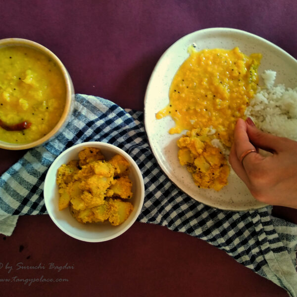 served plate of tok dal rice and aloo poshto with bowlfuls of tok dal and aloo poshto on checkered cloth and maroon background